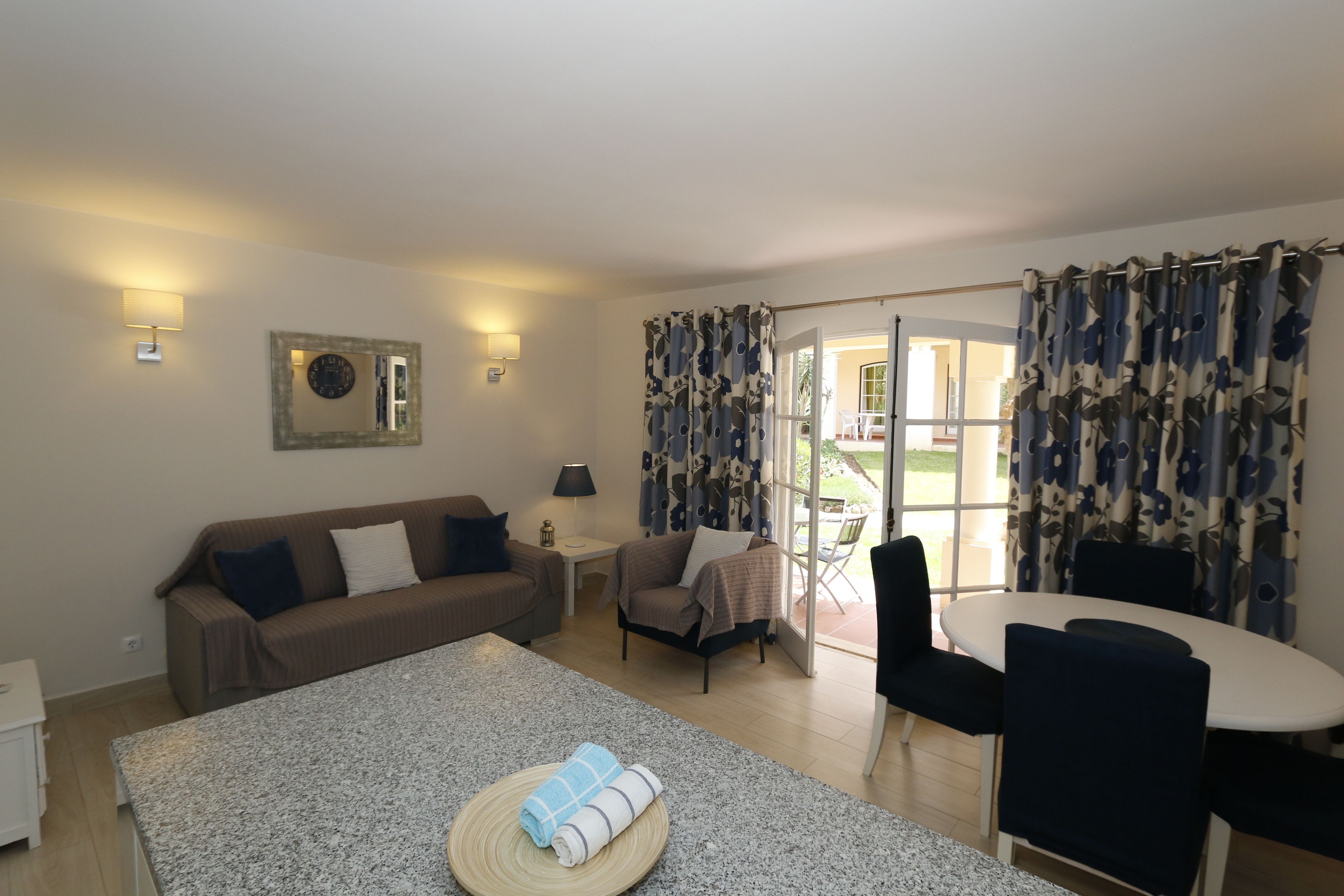 A room with a sofa, a table with a towel, a chair, a lamp, and a window with curtains.