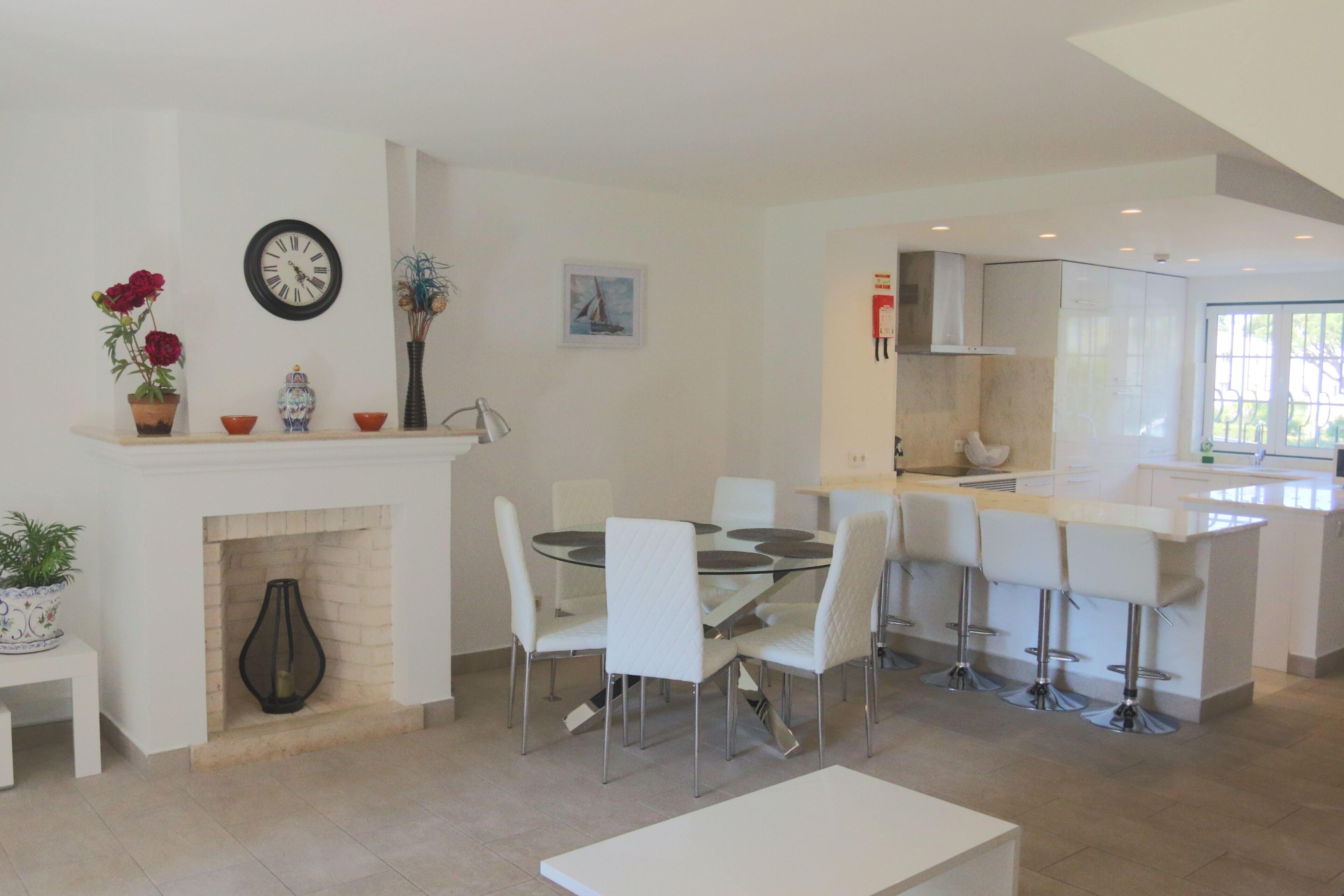 A modern kitchen with white cabinets, a dining area with a glass table and white chairs, and a fireplace with decorative items.