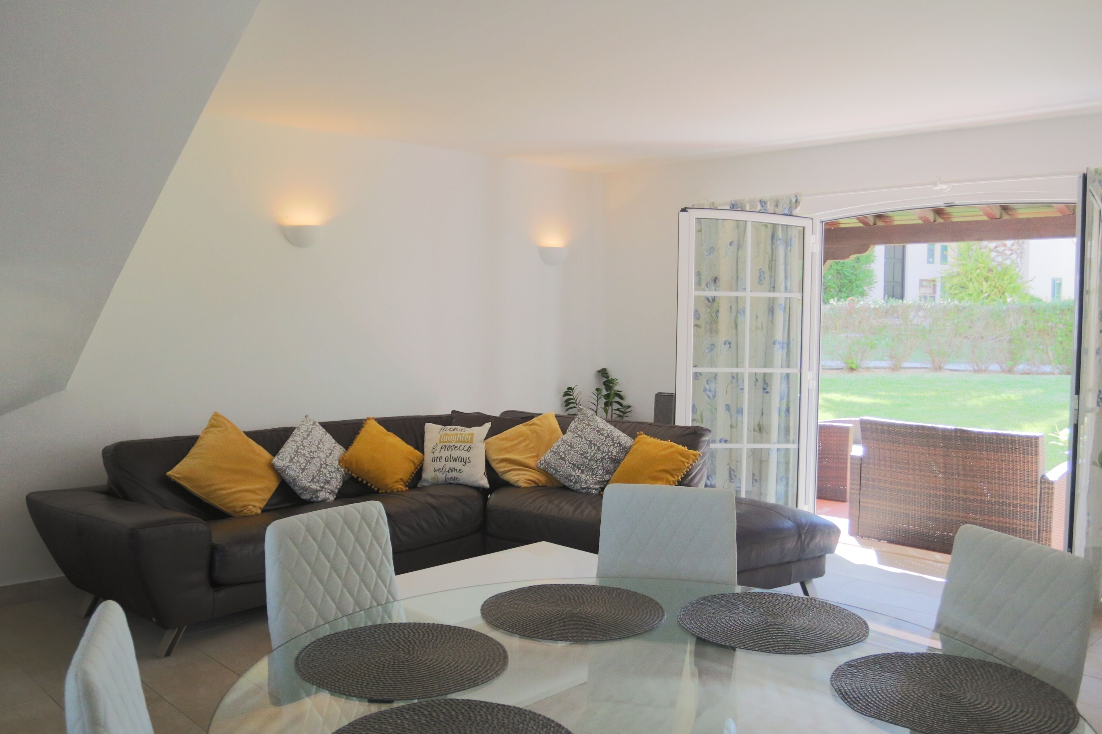 Ein modernes Wohnzimmer mit einem Glastisch, einem dunkelbraunen Ledersofa mit gelben Kissen und einer Glasschiebetür, die zu einem Außenbereich führt.