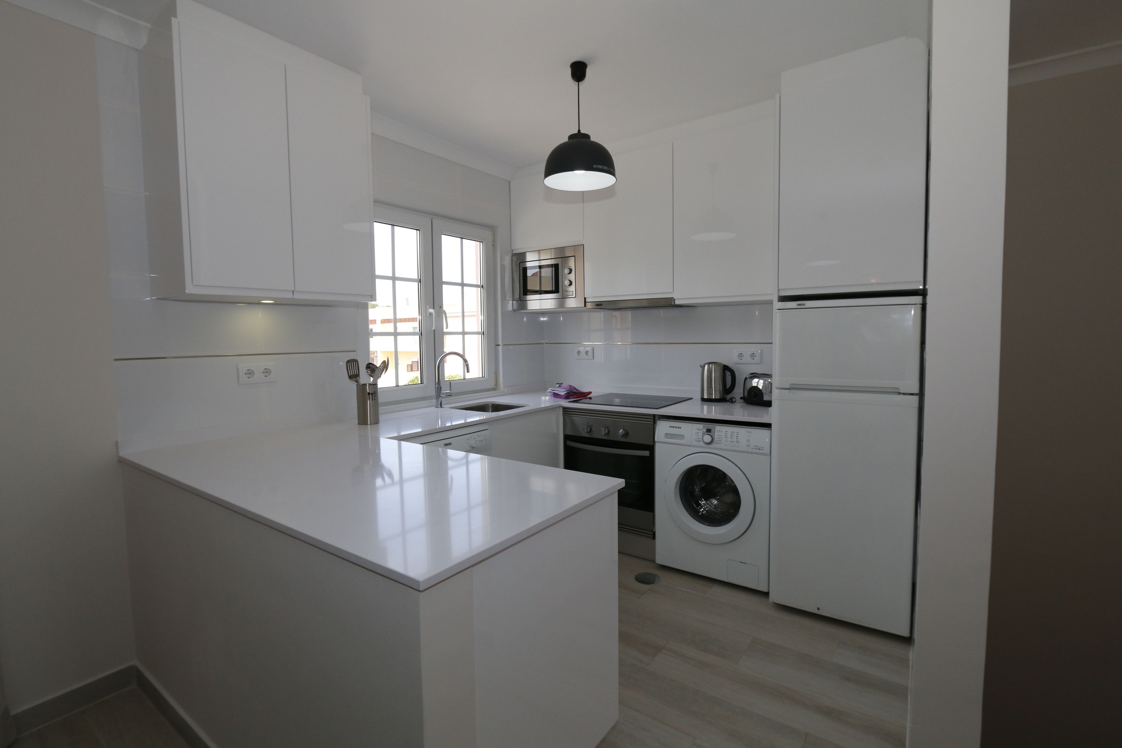 Une cuisine moderne avec des meubles blancs, un îlot central, un four encastré, un lave-linge et un réfrigérateur.