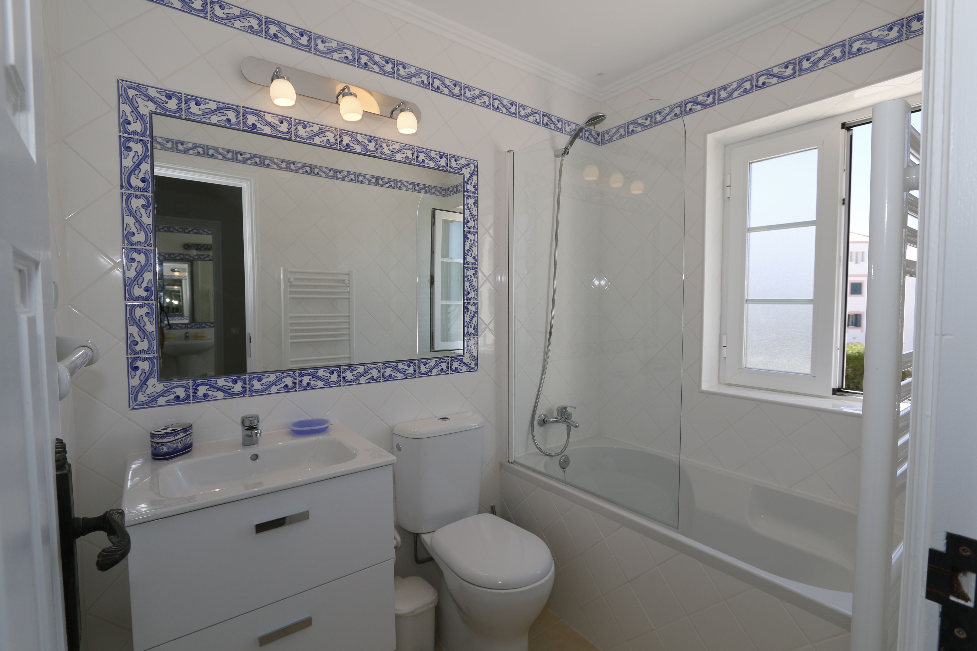 Un baño con lavamanos, inodoro y ducha blancos. Hay un espejo sobre el lavamanos con azulejos decorativos azules.