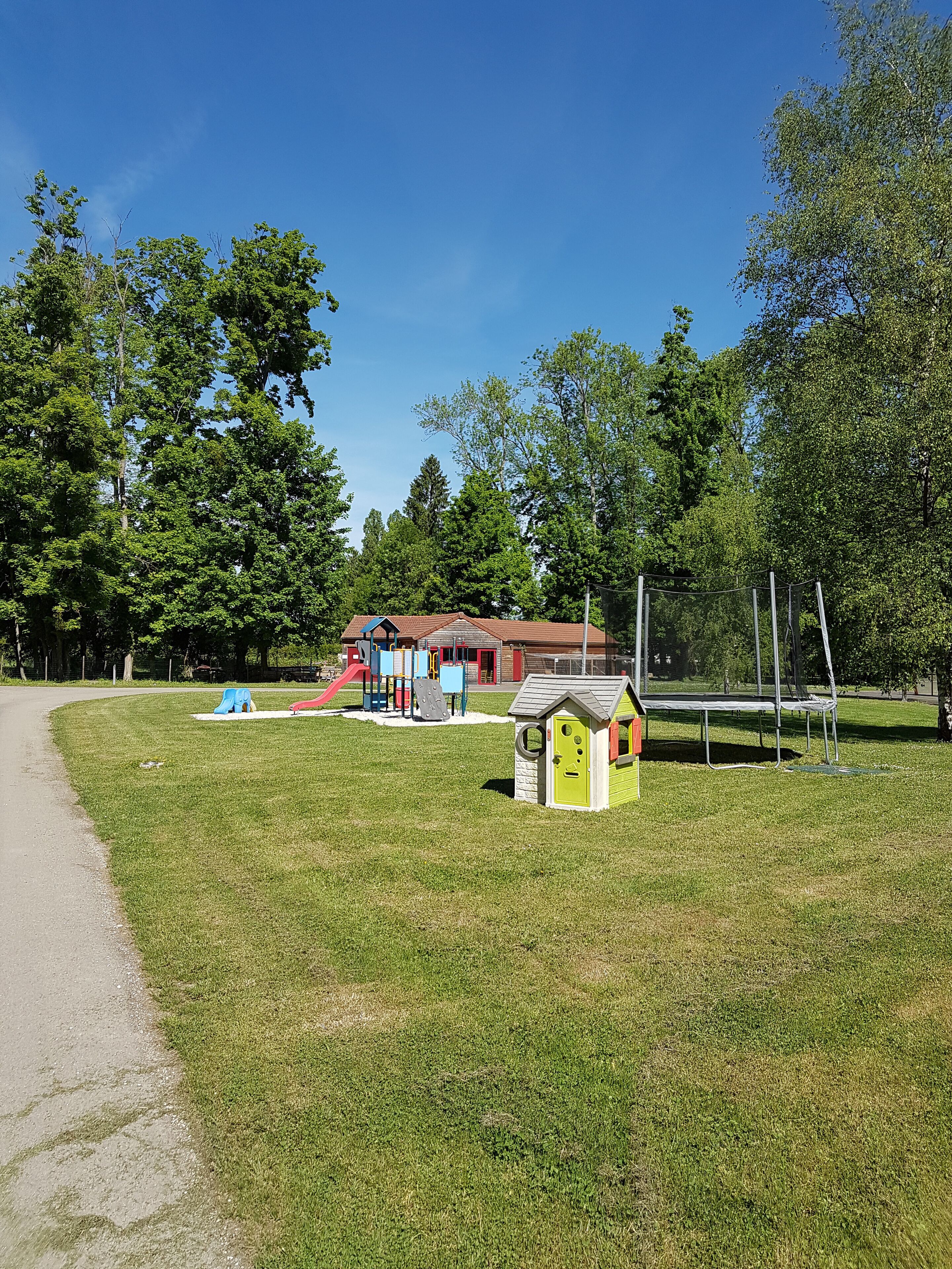 A playground with various play structures, a small green building, and a red building in the background.