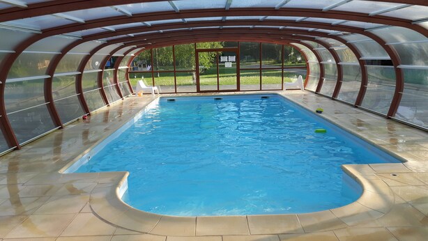 An indoor swimming pool with a glass ceiling, surrounded by a tiled floor and a wooden deck.