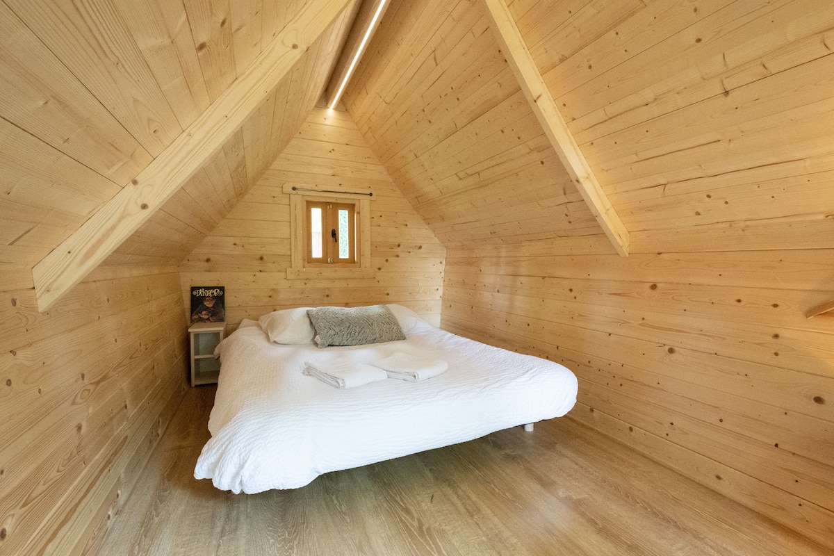 A cozy wooden room with a sloped ceiling, a bed with white bedding, a small bedside table, and a framed picture on the wall.