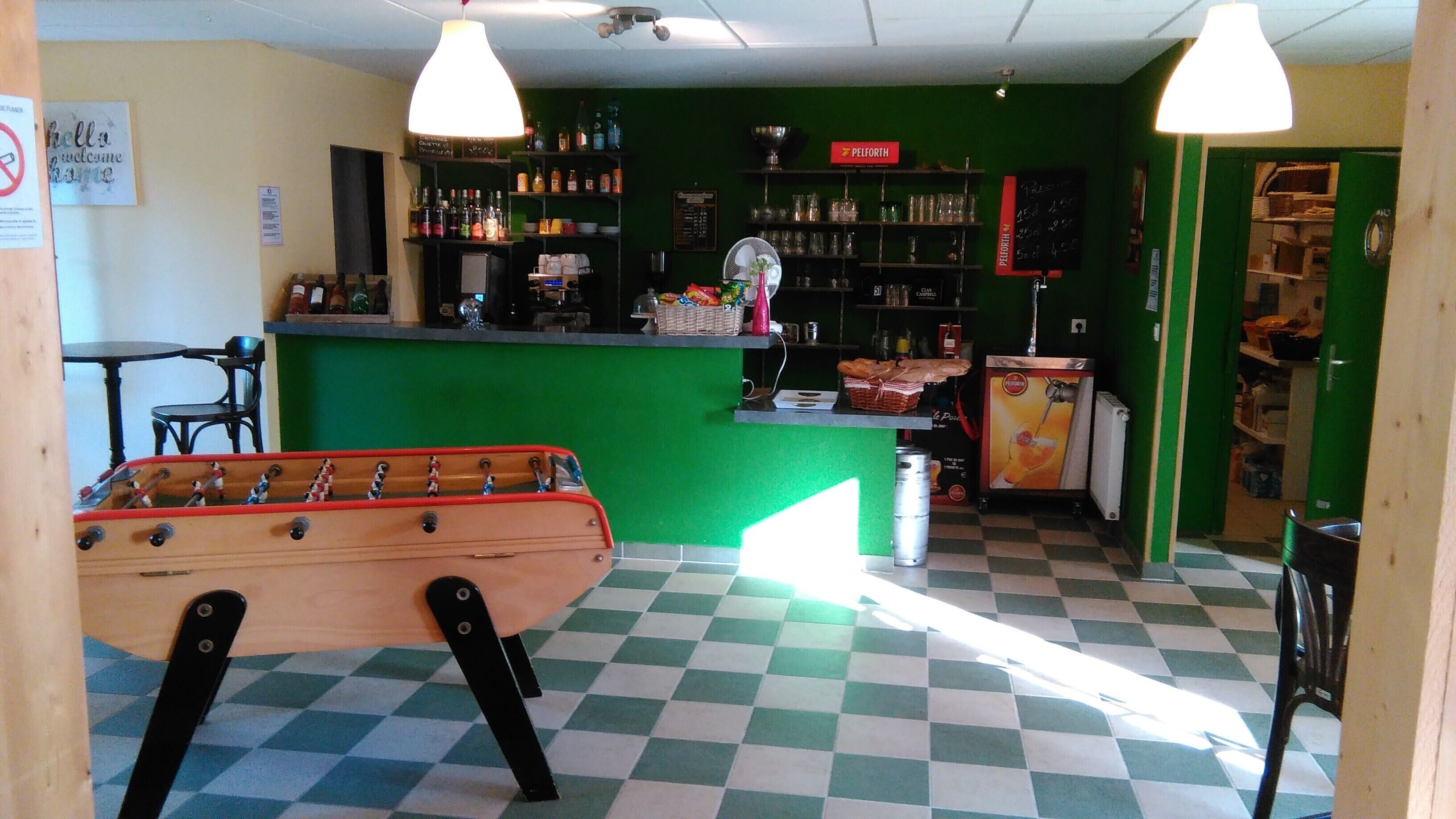 Zona de bar con una mesa de fútbol de mesa, un mostrador con varias botellas y una pared verde con estantes.