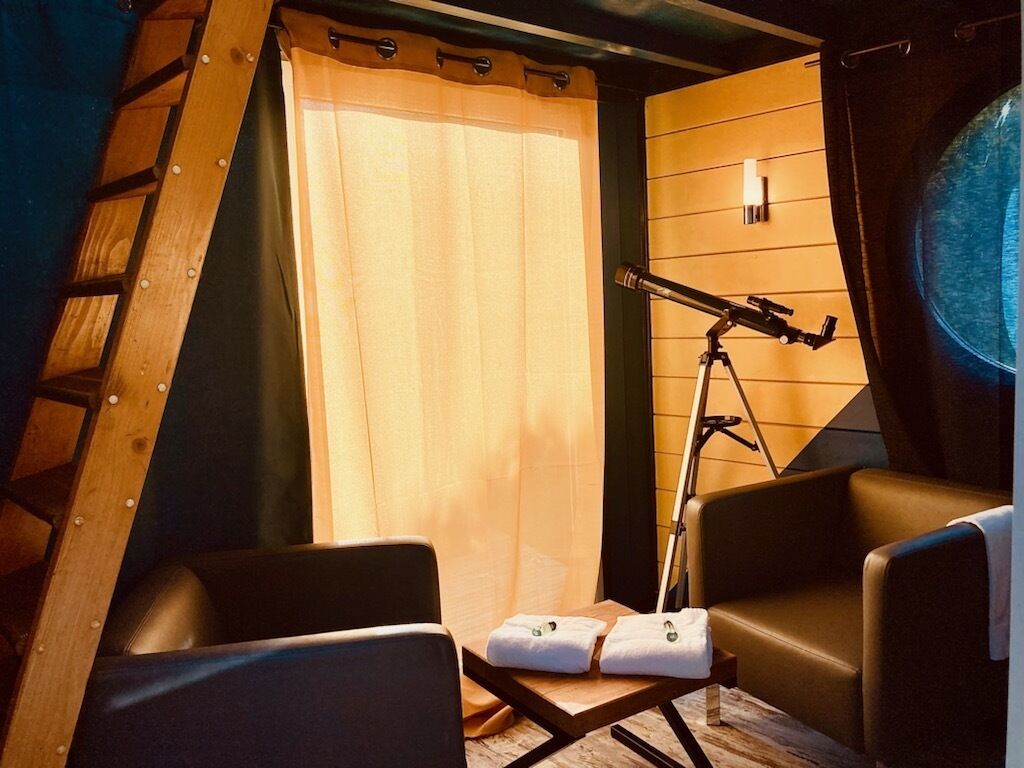 Een gezellige kamer met een telescoop, twee stoelen, een klein tafeltje met handdoeken, en een raam met gordijnen.