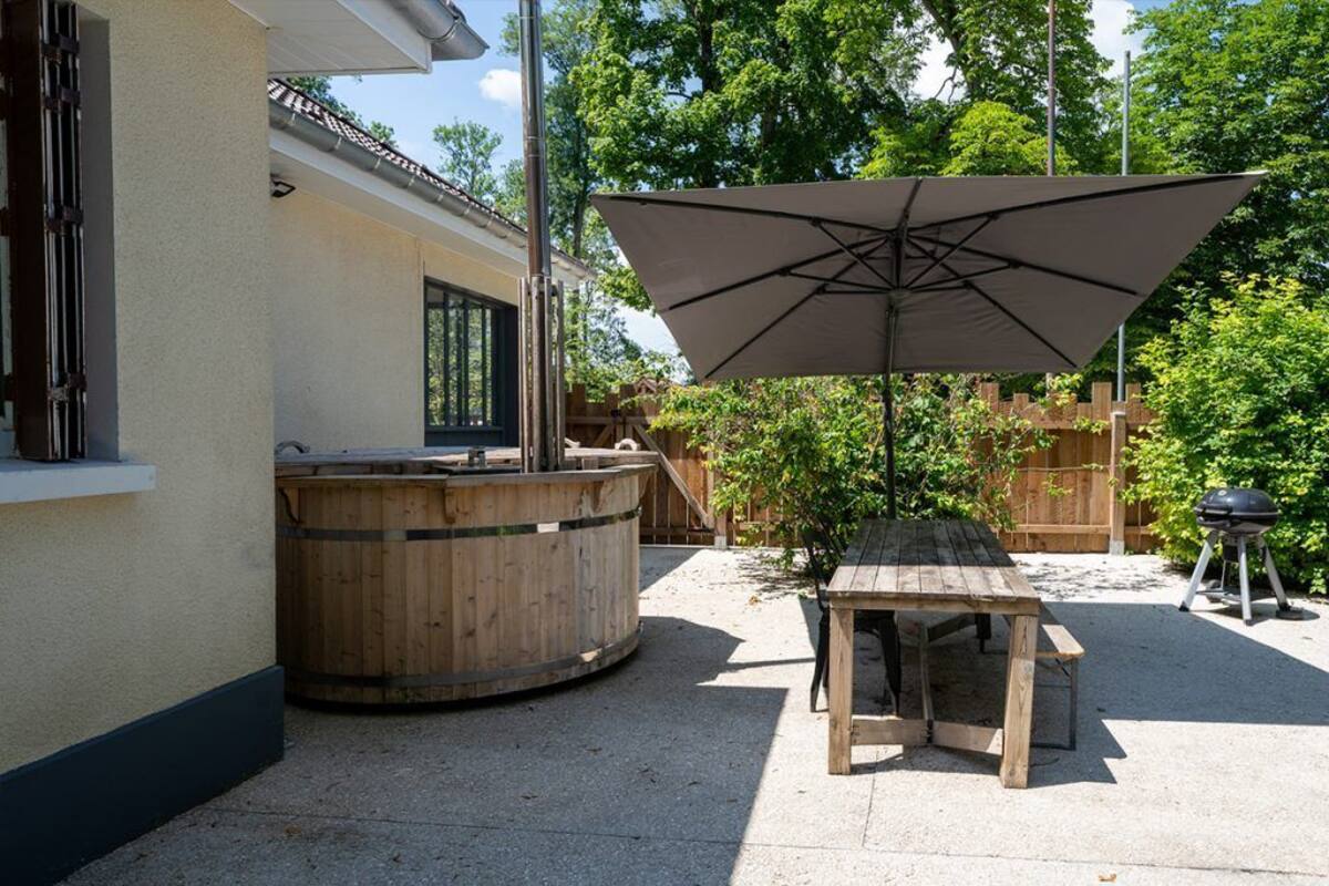 An outdoor patio with a wooden table and a large umbrella, a wooden hot tub, and a barbecue grill.