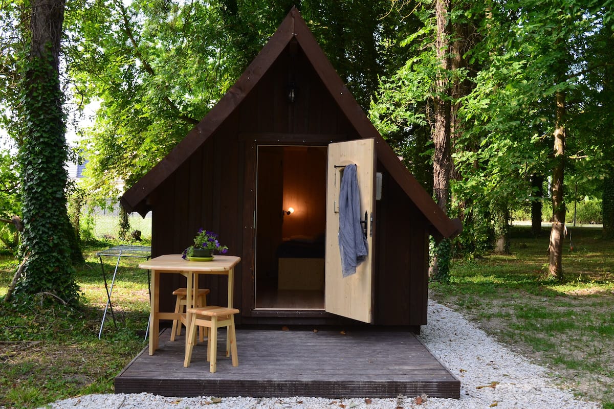 A small wooden cabin with an open door, a table with chairs, and a garden setting.