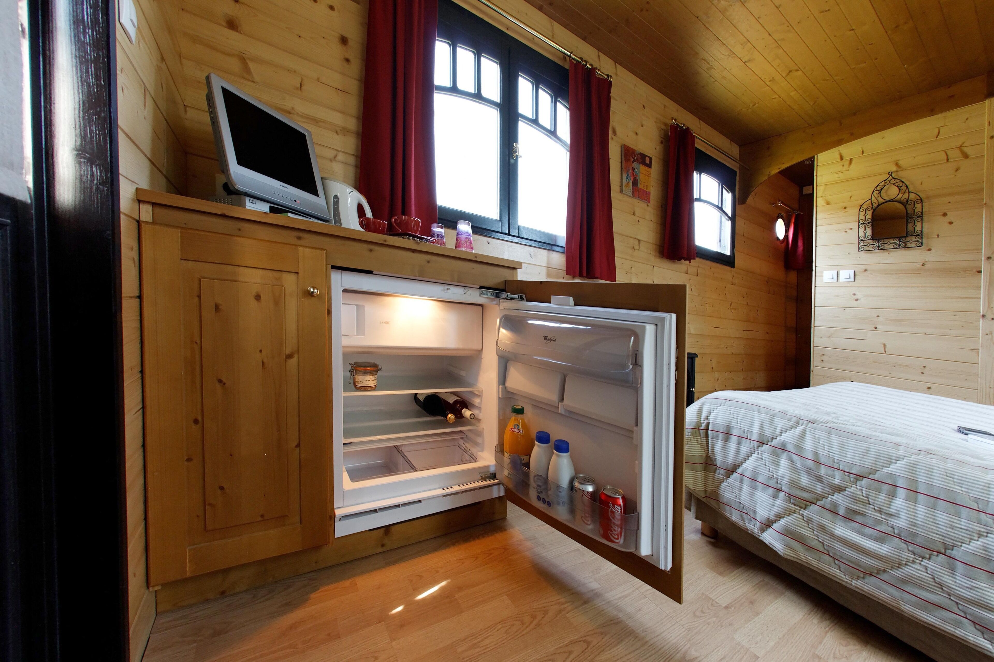 A wooden cabin room with a bed, a built-in TV, a refrigerator, and a window with curtains.