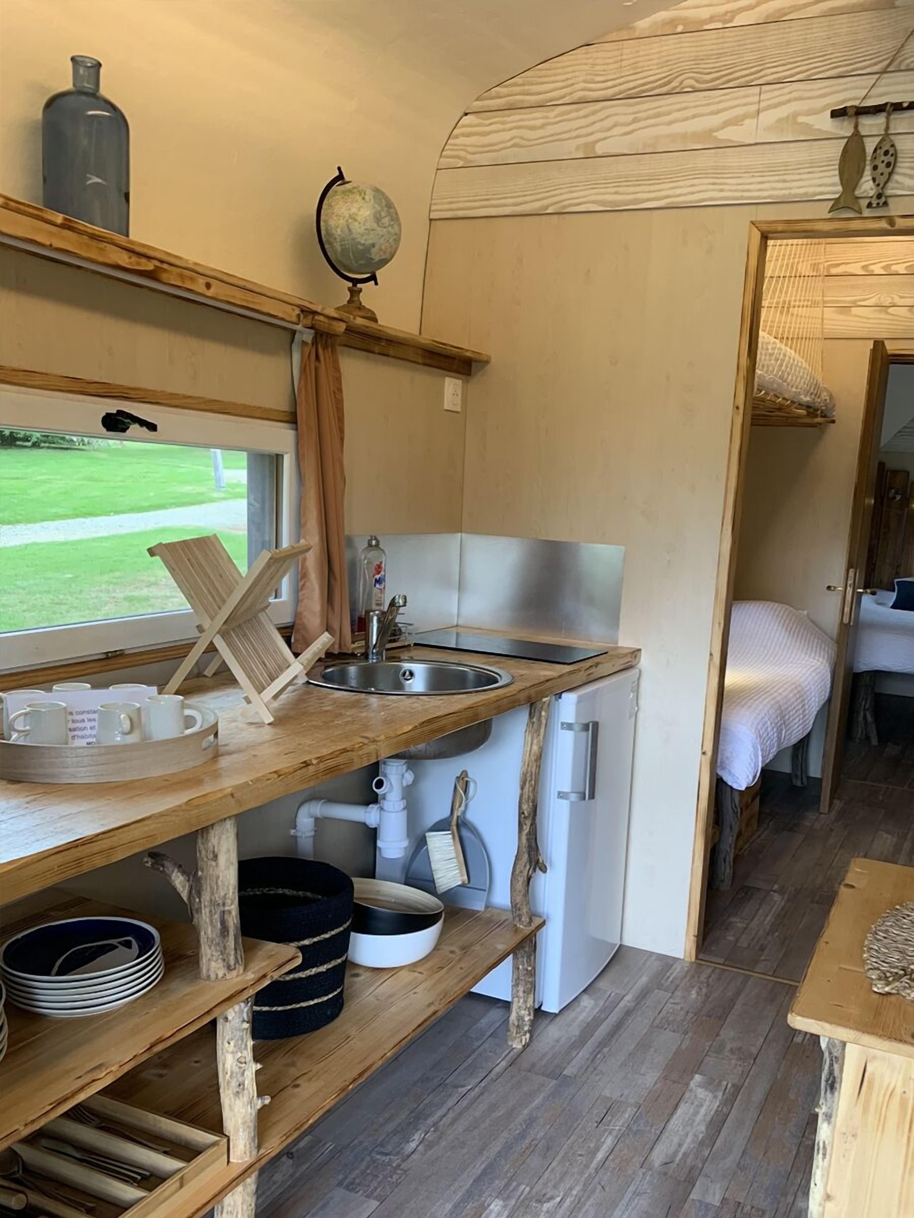 Cocina compacta con un lavamanos, un refrigerador pequeño y estantes de madera con utensilios de cocina.