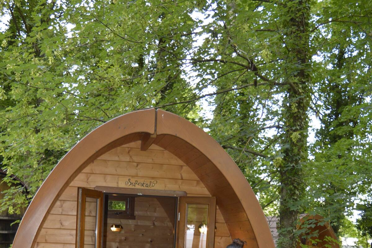 A wooden arched structure with a bed inside, a person sitting at a table with a chair, and a small table with a candle.