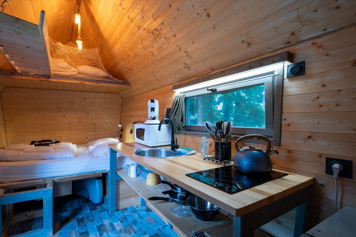 A compact wooden cabin with a kitchenette, a bed with white linens, a window with a view of trees, and a hanging light fixture.