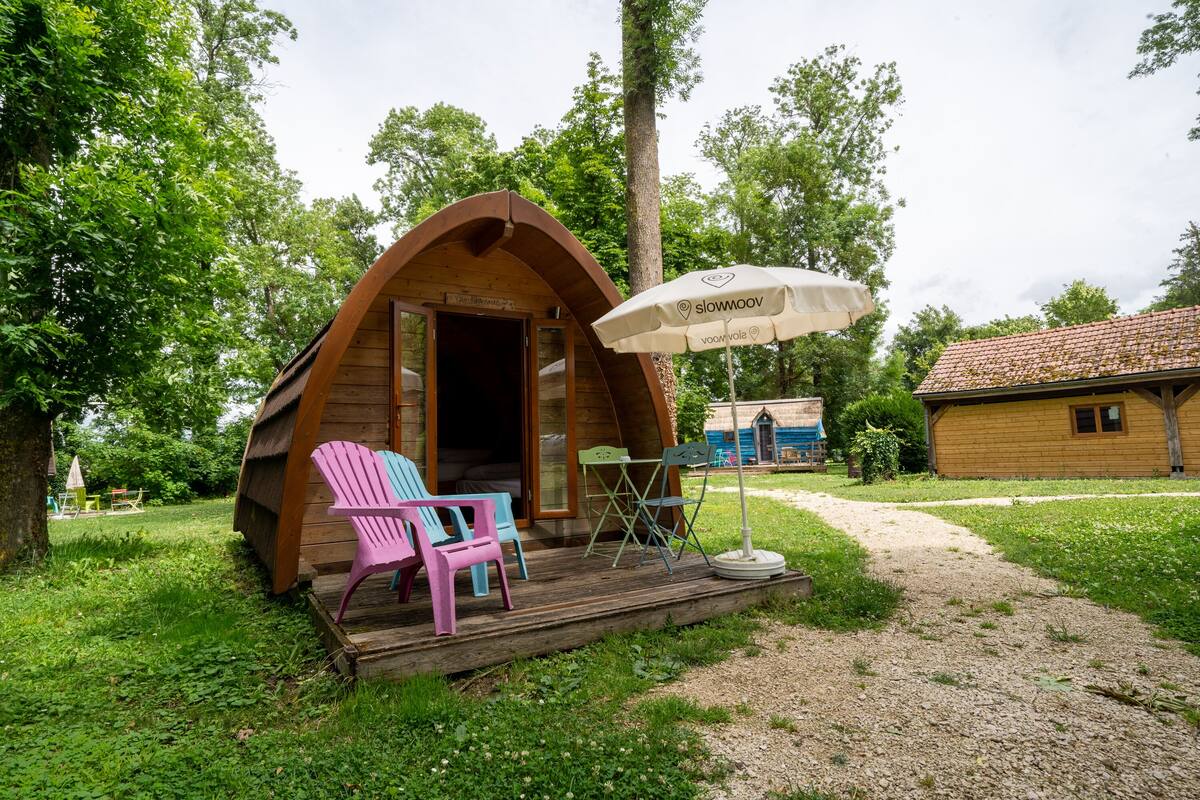 A wooden cabin with an open door, a small porch with colorful Adirondack chairs, and a table with a white umbrella.