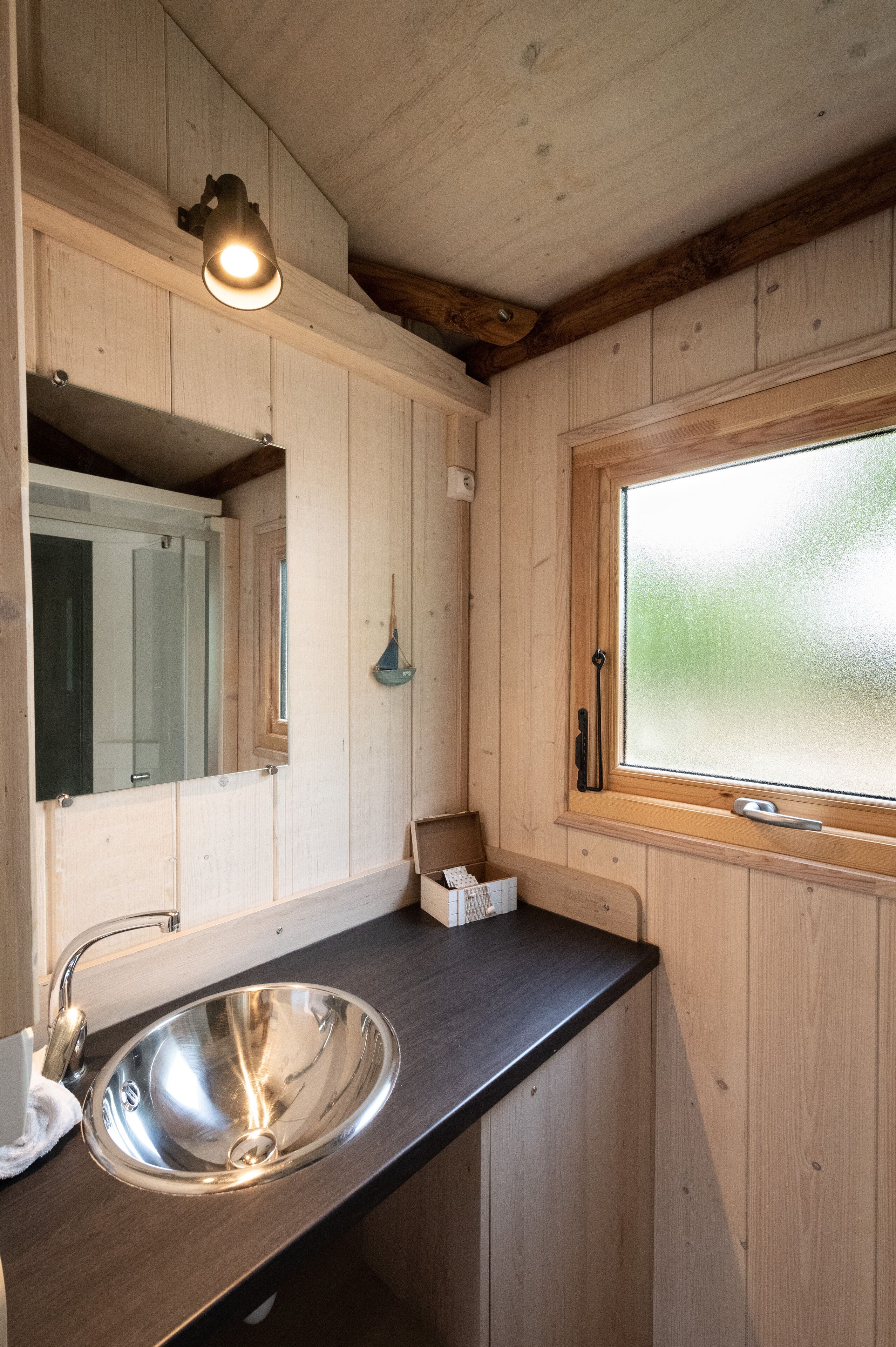 A wooden cabin interior with a metal sink, a mirror, a window with frosted glass, and a hanging light fixture.