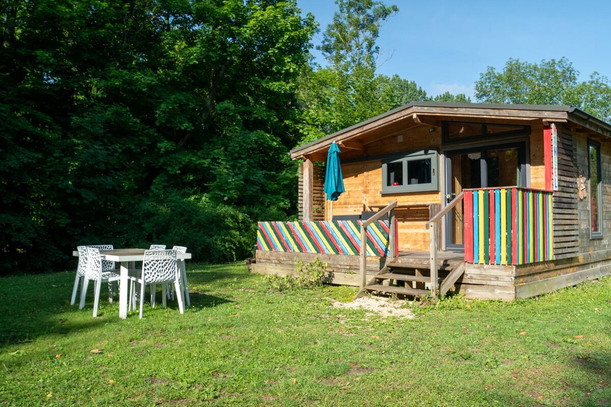 A wooden cabin with a colorful striped awning, a small porch, and a table and chairs set outside.