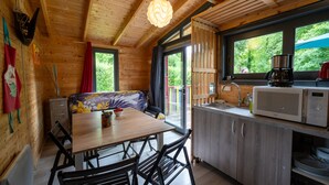 A wooden cabin with a dining table, chairs, a bench, a kitchen area with a microwave, and a window overlooking greenery.