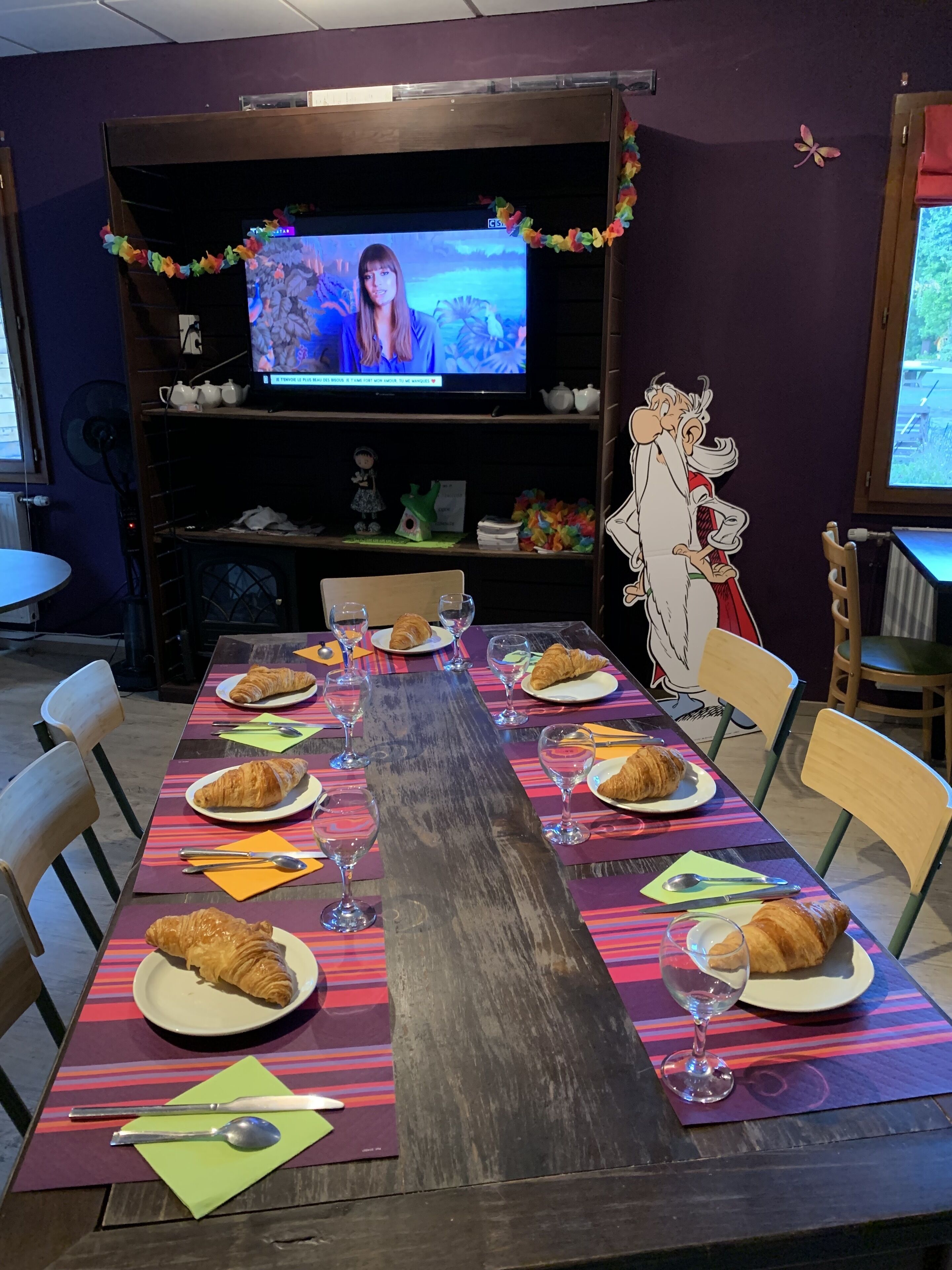 A long dining table set with croissants, wine glasses, and plates, with a television in the background.