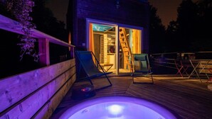 A wooden deck with a hot tub, two lounge chairs, and a sliding glass door leading to a room.