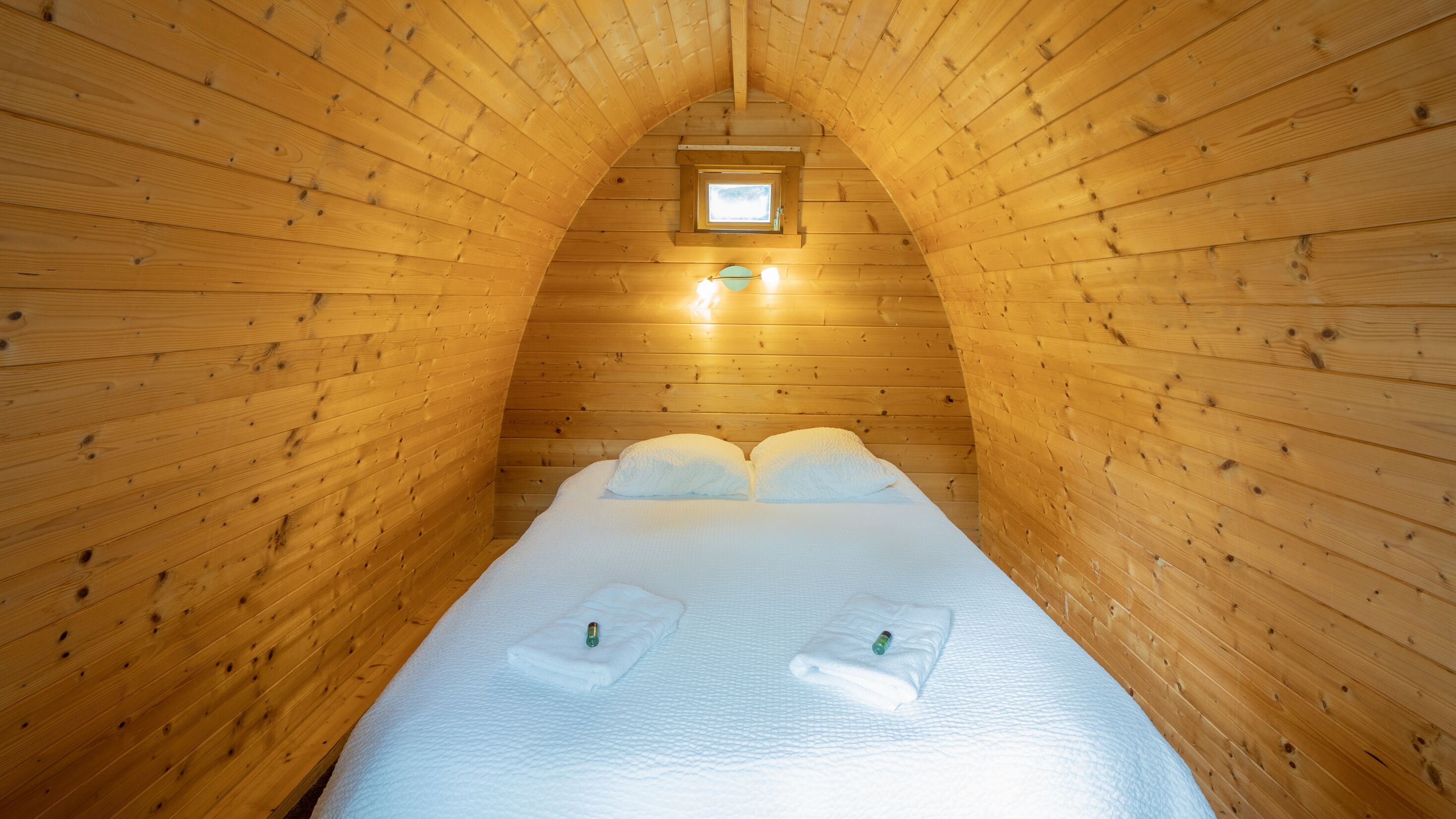 Une pièce étroite aux murs lambrissés, avec un lit placé au centre, deux tables de chevet, une petite fenêtre et un luminaire au-dessus du lit.