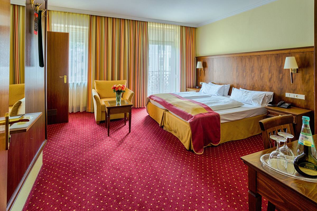 A hotel room with a large bed, a desk with a telephone, a chair, a small table with a vase of flowers, and a red carpeted floor.