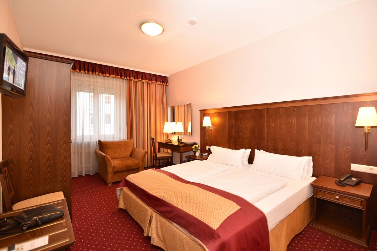 A hotel room with a large bed, a desk with a lamp, a chair, and a wooden wardrobe.