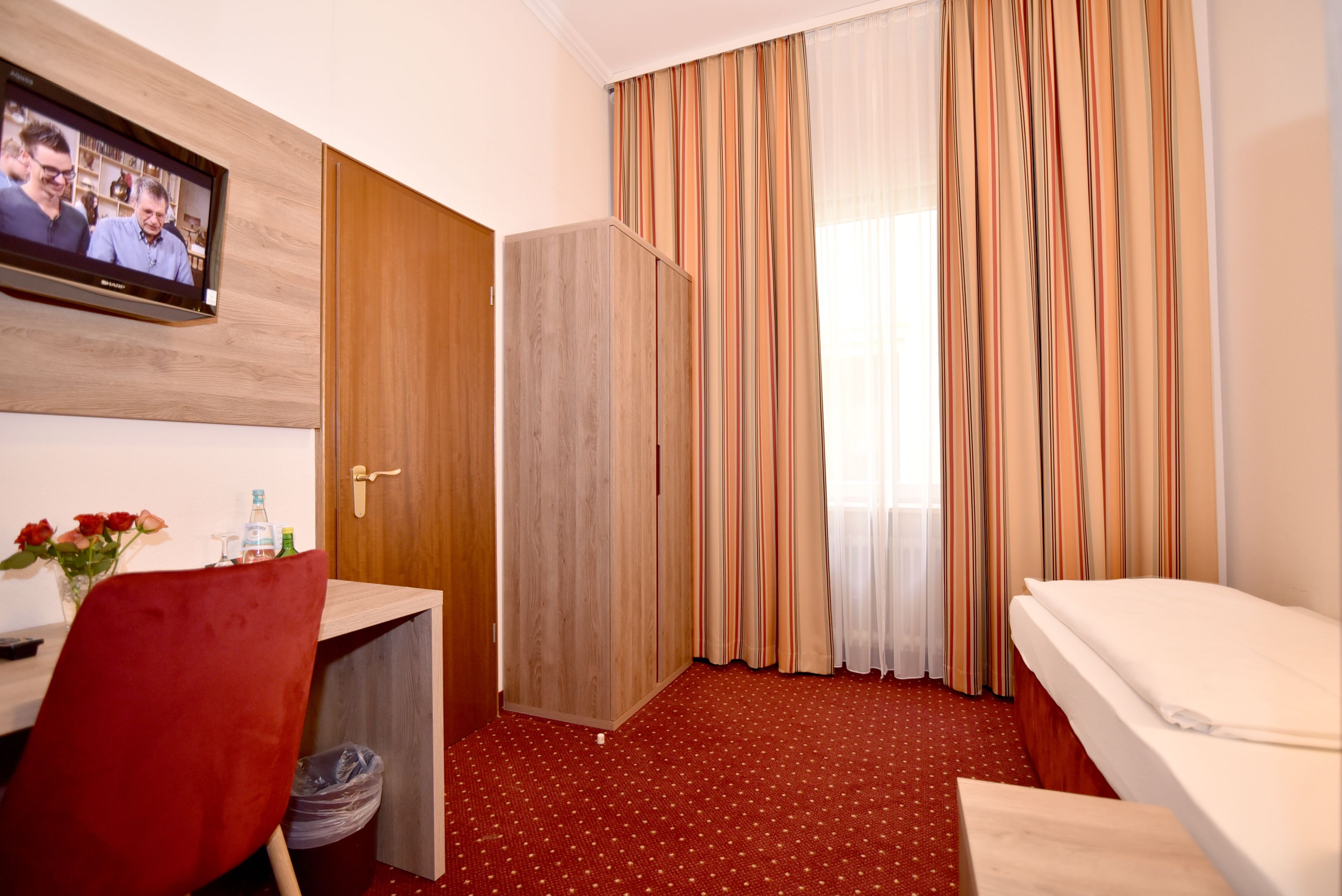 A hotel room with a wooden wardrobe, a desk with a red chair, a bed, and a TV mounted on the wall.