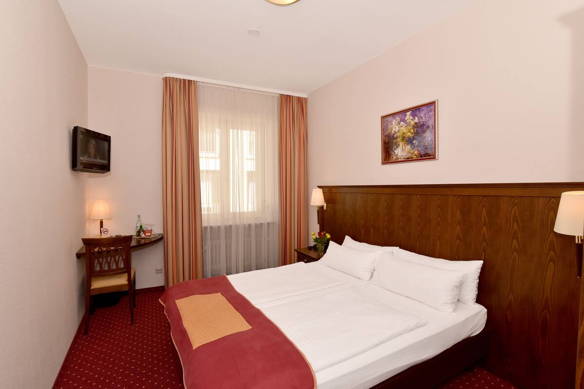 A hotel room with a large bed, a desk with a chair, a television mounted on the wall, and a painting above the bed.