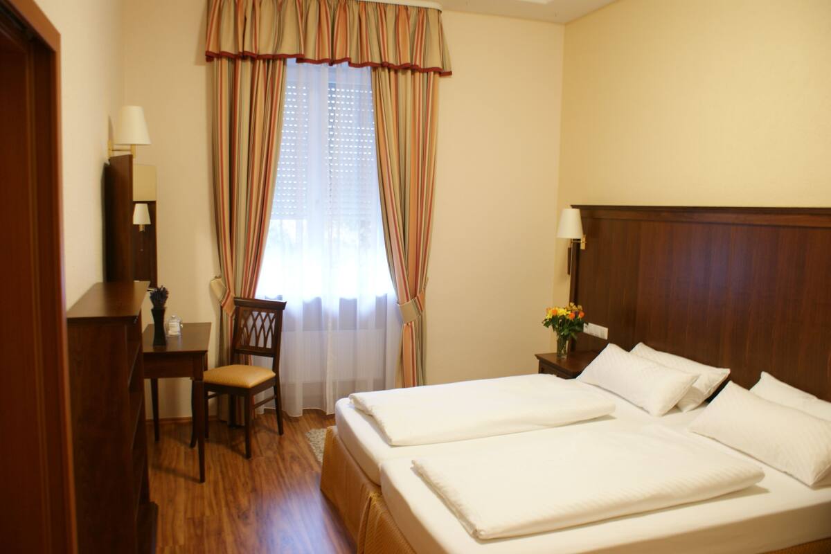 A hotel room with two beds, a desk, a chair, a lamp, and a window with curtains.