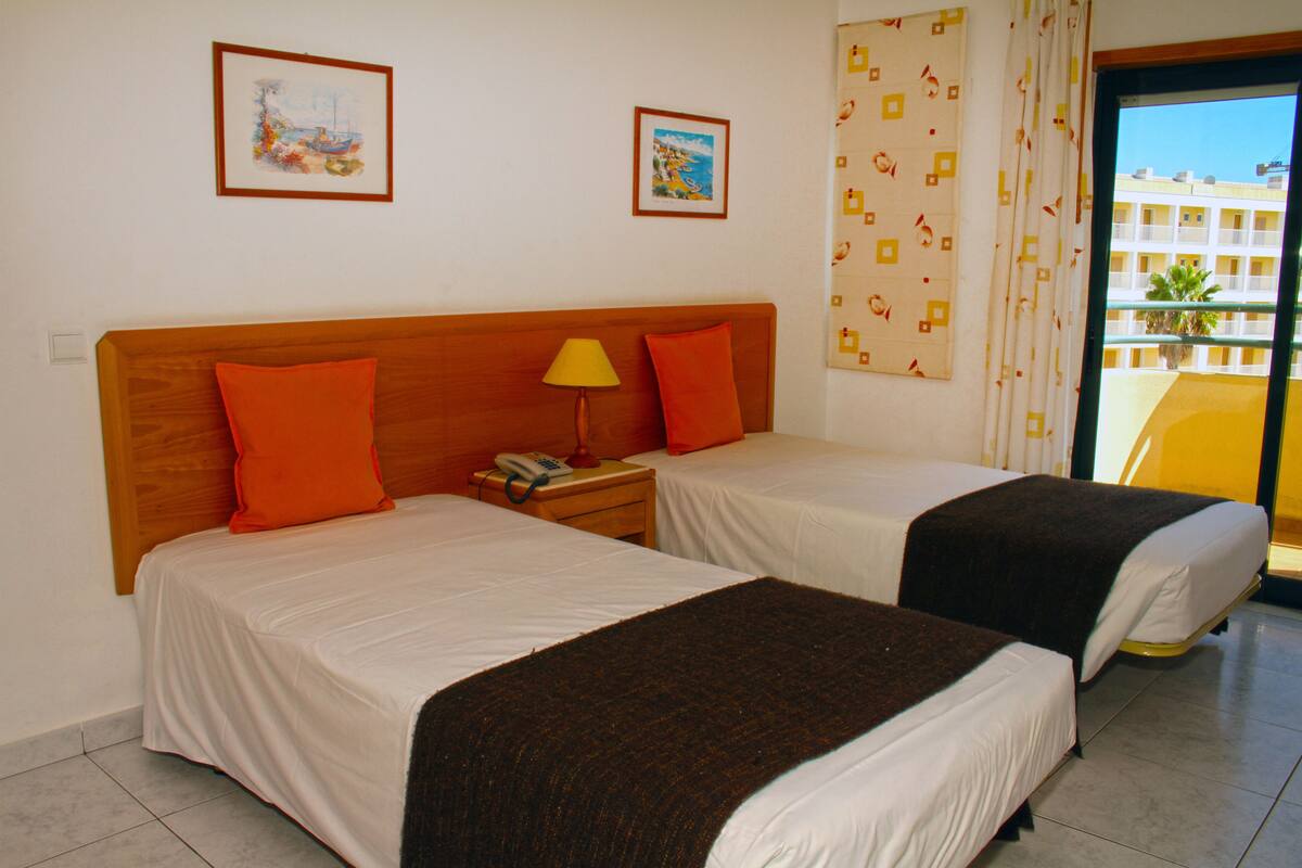 Une chambre d’hôtel avec deux lits, une tête de lit en bois, une table de chevet avec une lampe et deux tableaux encadrés au mur.