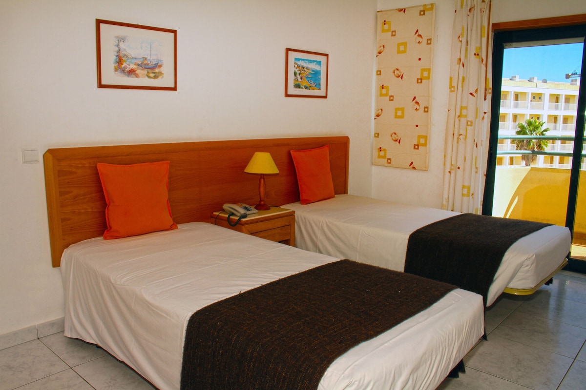 A hotel room with two beds, a nightstand with a lamp, and a view of a building through a window.