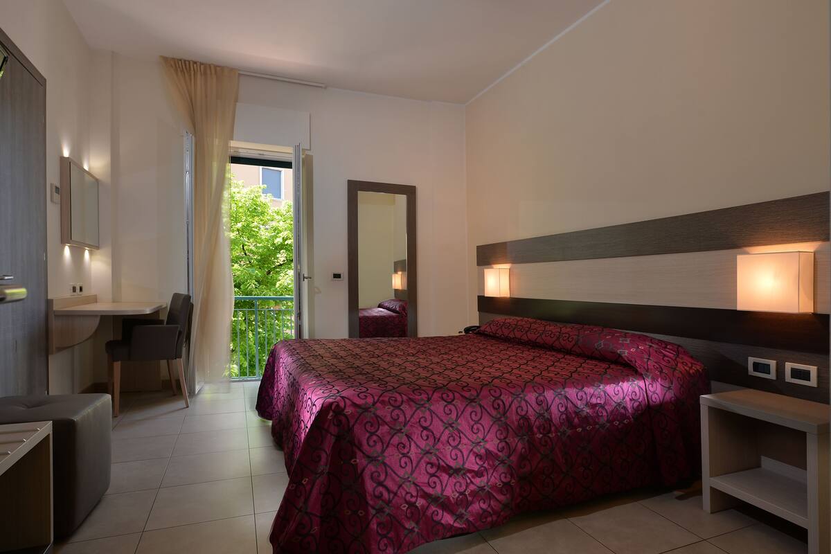 A bedroom with a large bed, a desk, a chair, and a balcony with a view of greenery.