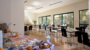 A spacious dining area with multiple tables and chairs, large windows offering natural light, and a buffet with various food items. - Hotel Siena (Verona)