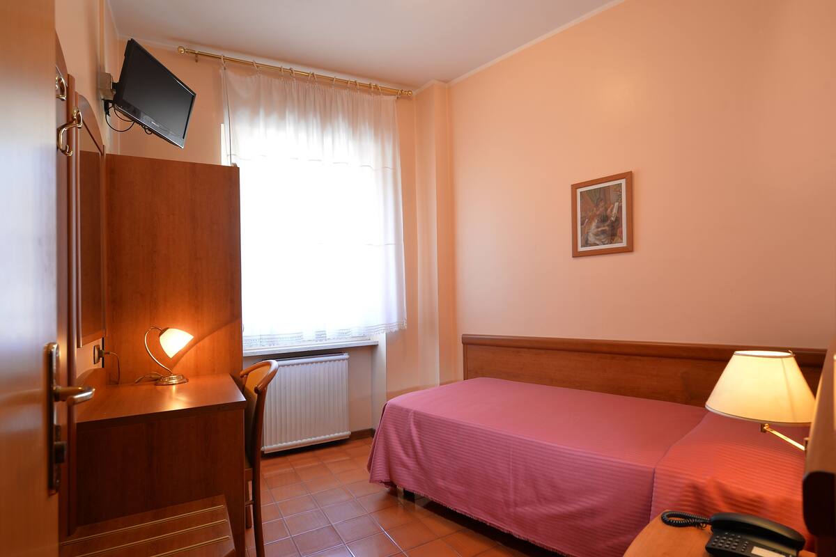 A hotel room with a bed, a desk with a lamp, a telephone, a picture on the wall, and a window with curtains.