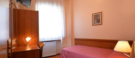 A hotel room with a bed, a desk with a lamp, a telephone, a picture on the wall, and a window with curtains.
