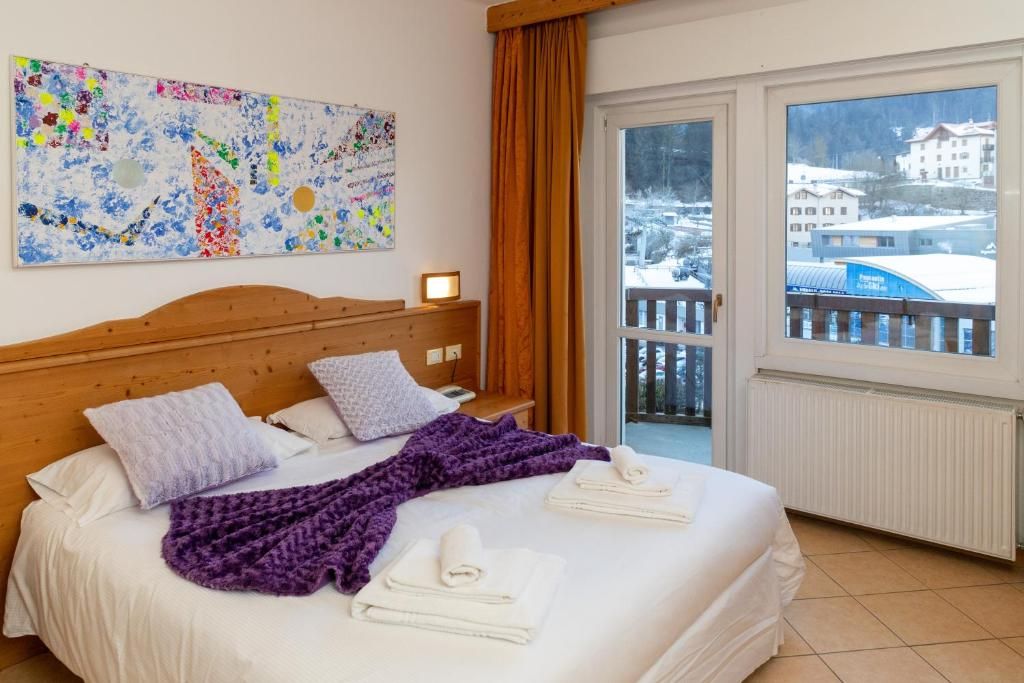 Una camera d'albergo con un letto, una finestra vista su un paesaggio innevato, una testiera in legno e un quadro astratto dai colori vivaci appeso al muro.