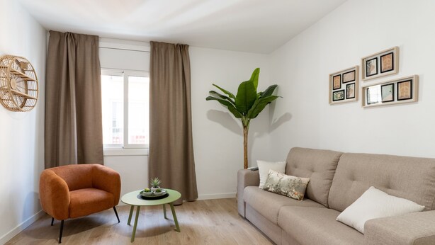 Een moderne woonkamer met een beige bank, een oranje fauteuil, een klein rond tafeltje met een plant en een grote groene plant naast de bank.