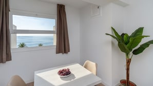 Un coin repas avec une table, un saladier de fruits, une plante en pot et une vue sur la mer par la fenêtre.