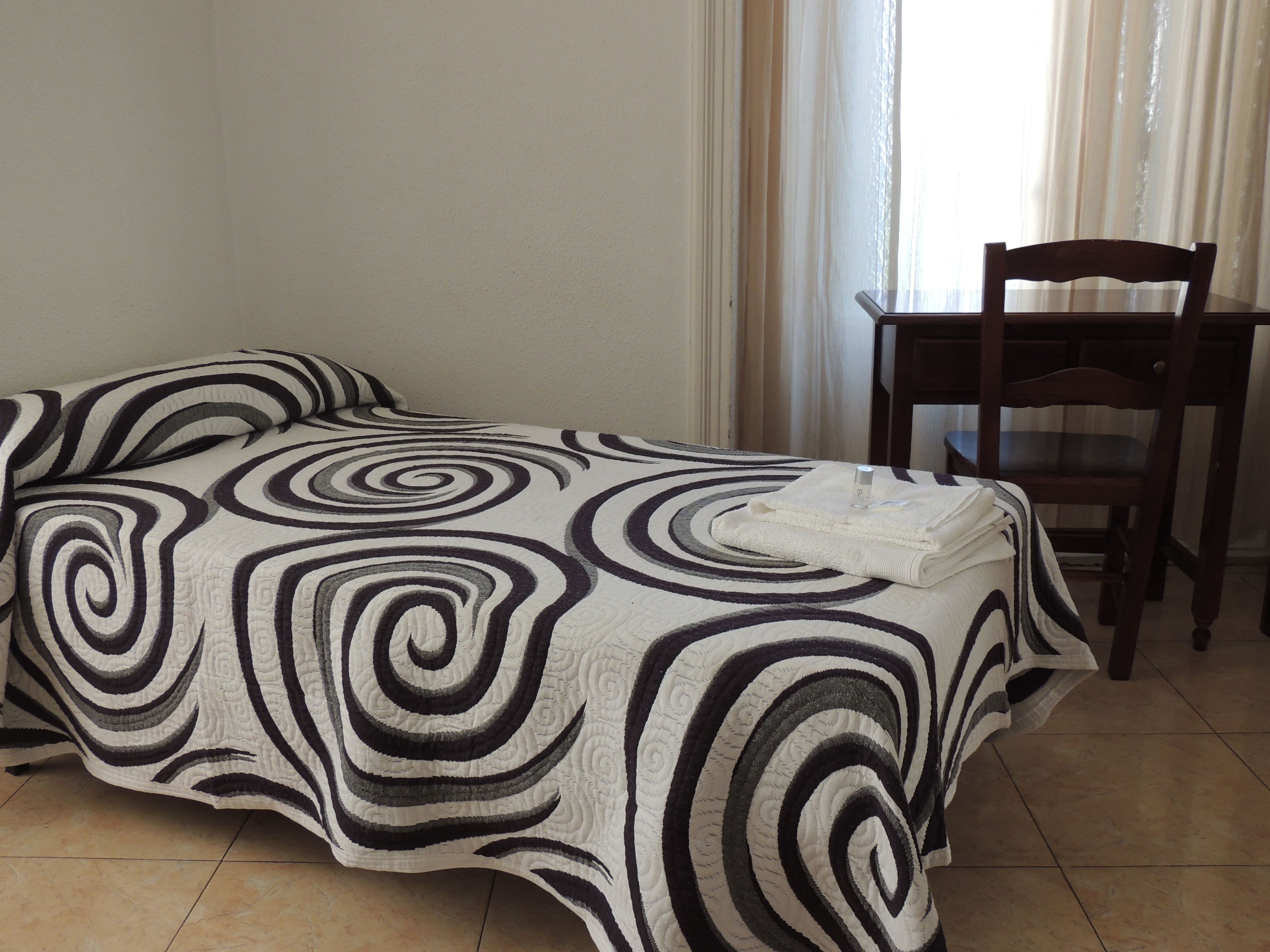 Una cama con una colcha de espirales blanco y negro, una toalla blanca doblada sobre la cama y una silla de madera al lado.