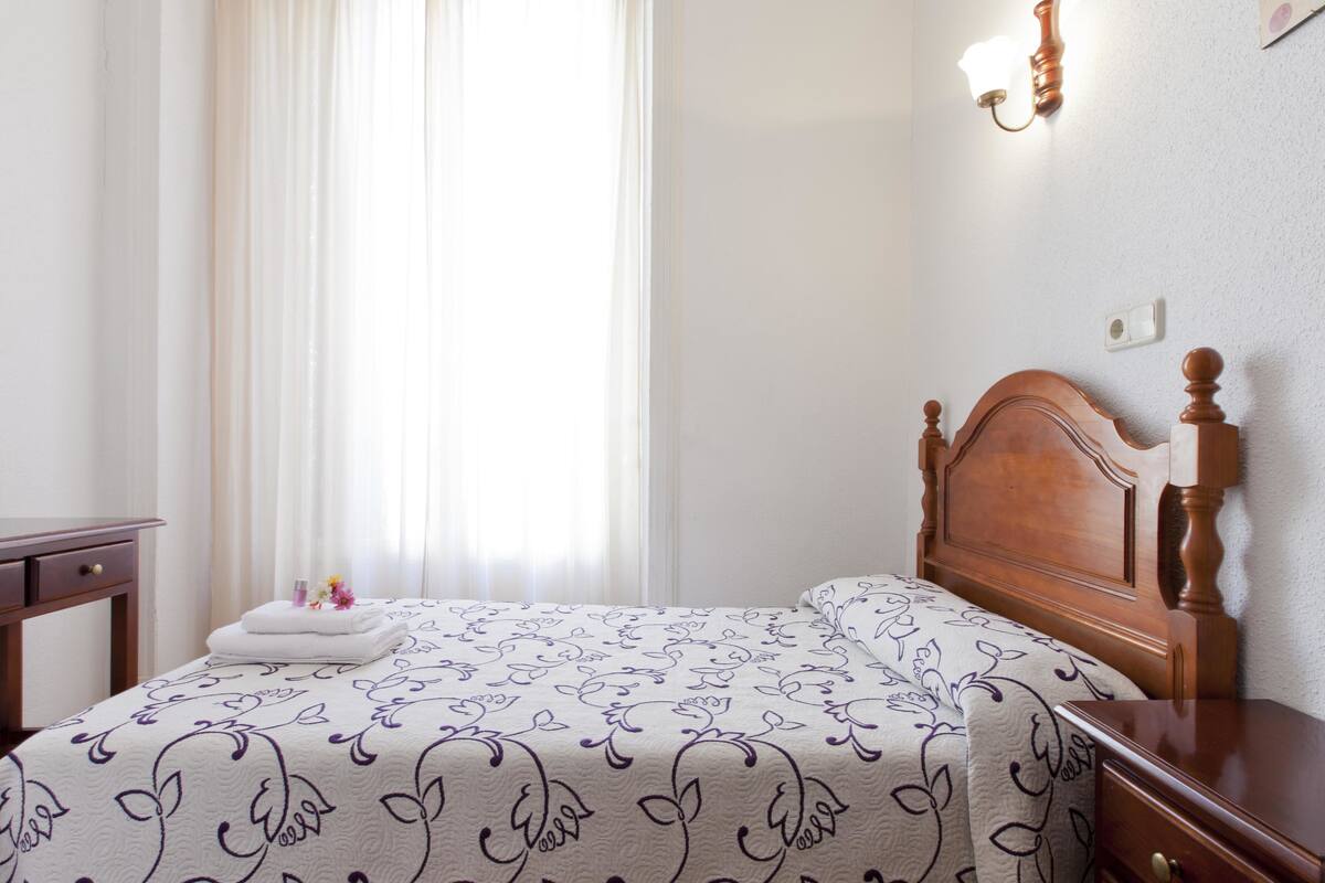 A neatly made bed with a floral-patterned bedspread, a wooden headboard, and a bedside table with a lamp.