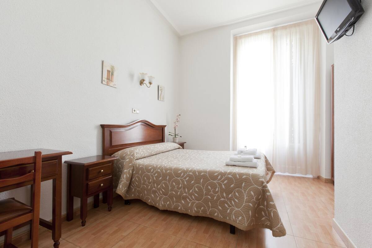 A neatly arranged bedroom with a bed, bedside tables, a desk, and a television mounted on the wall.