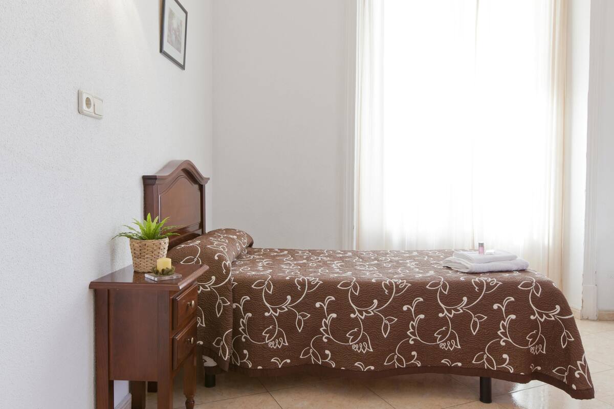 A neatly made bed with a patterned bedspread, a bedside table with a plant, and a window with curtains.