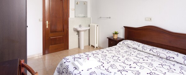 Un dormitorio con puerta de madera, lavamanos blanco, radiador, mesita de noche con planta y cama con colcha de motivos florales.