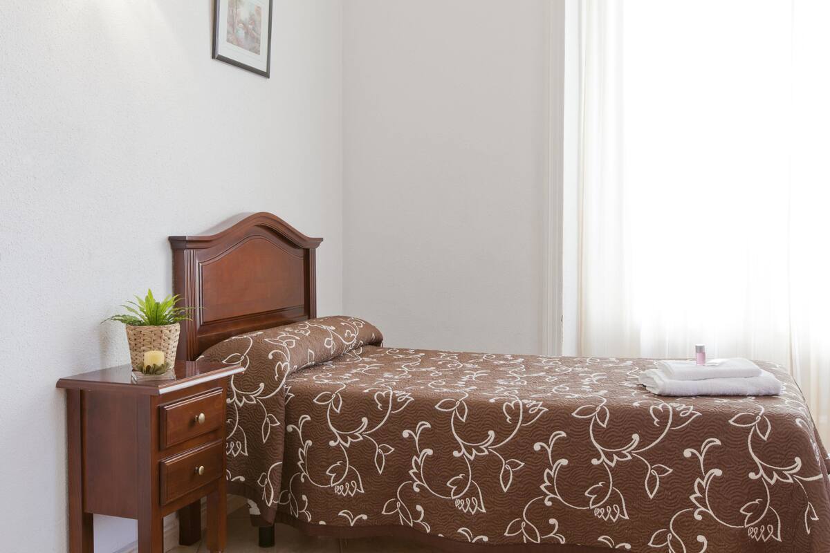 A neatly made bed with a brown patterned bedspread, a wooden bedside table with a plant, and a framed picture on the wall.