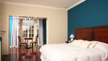 Un dormitorio con una cama grande, una mesita de noche de madera, una mesa con sillas y vistas a través de una ventana con cortinas.