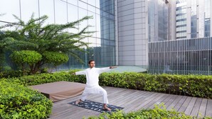 Uma pessoa praticando yoga em um deck de madeira, com vista para edifícios modernos e vegetação.