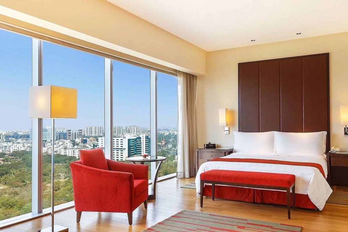 Une chambre d’hôtel moderne avec un grand lit, un fauteuil rouge, une petite table et une vue sur la ville grâce à de grandes fenêtres.