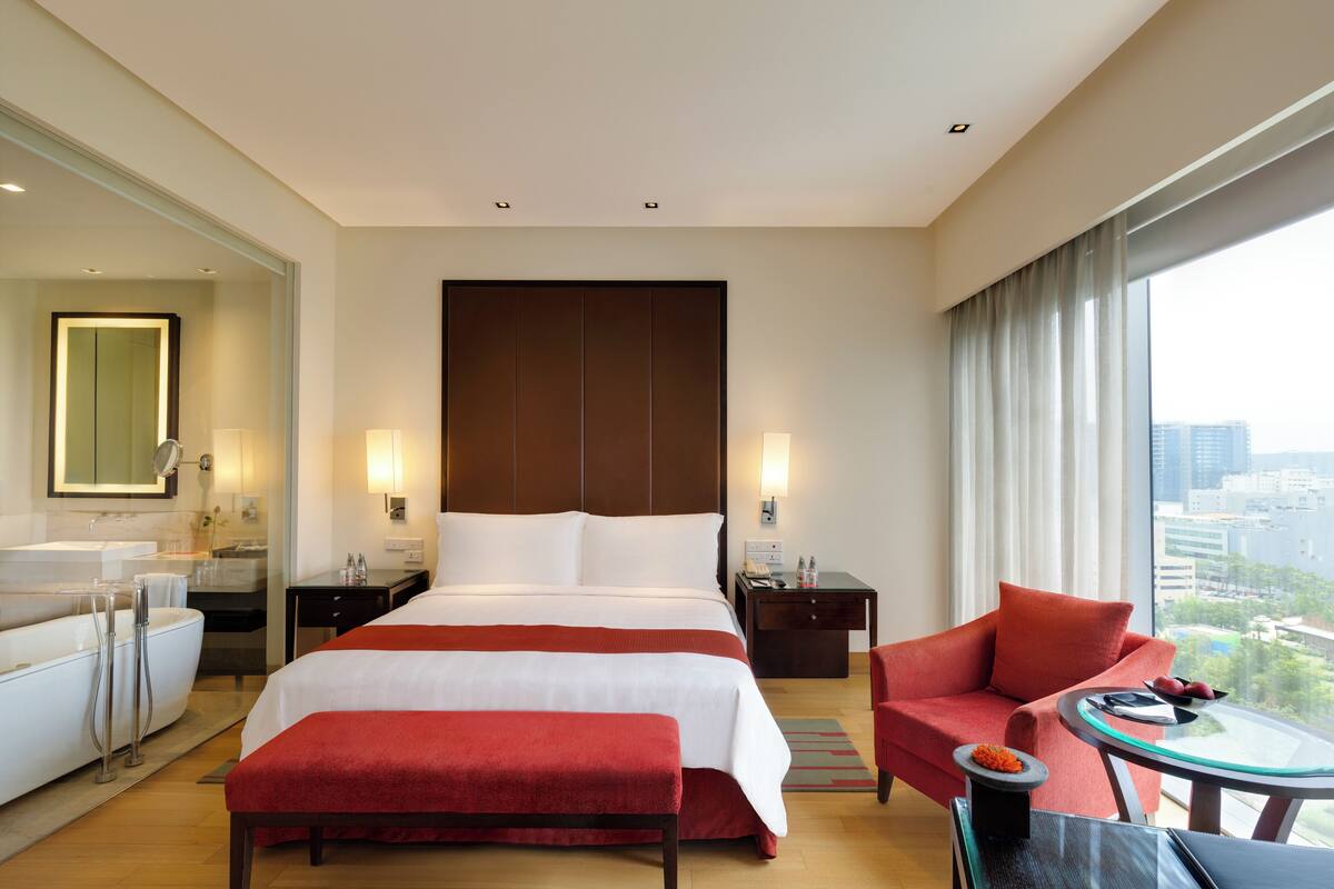 Une chambre d’hôtel avec un grand lit, un coin salon comprenant un fauteuil rouge et une table en verre, et une vue sur la ville grâce à de grandes fenêtres.