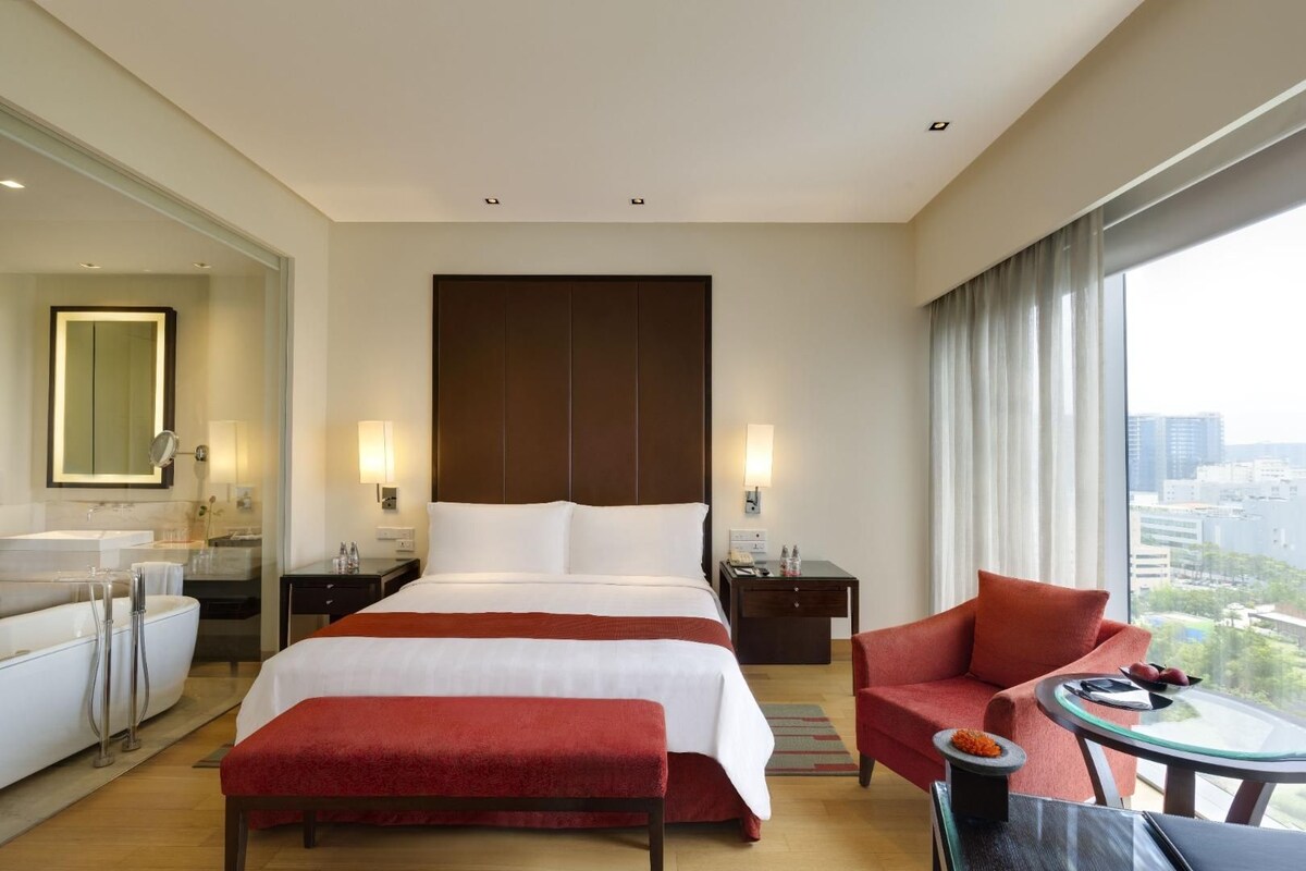 A hotel room with a large bed, a sitting area with a red chair, a small table, a sofa, a mirror, and a view of the city.