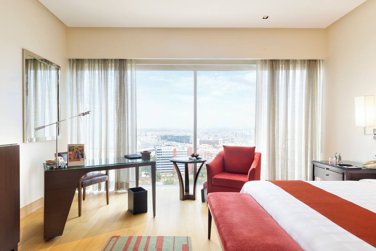 Une chambre d’hôtel avec un grand lit, un bureau, une chaise, une petite table et une vue sur la ville grâce à de grandes fenêtres.
