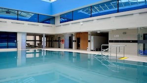 Una piscina interior con techo alto y tragaluces, rodeada de sillones de descanso y un flotador.
